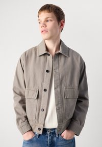 Ung mann med hendene i lommene iført beige jakke over hvit ribbestrikket skjorte og blå jeans, ser sidelengs mot ensfarget bakgrunn.