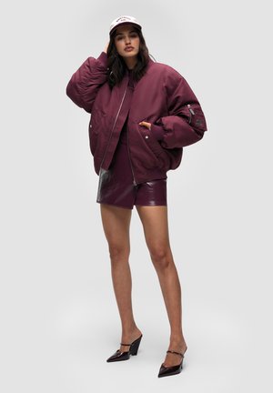 Bordowa kurtka bomber z prążkowanymi mankietami, w zestawie z błyszczącą bordową mini spódniczką. Całość uzupełniają czarne szpiczaste szpilki i jasna czapka.
