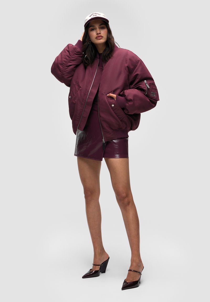 Vinröd bomberjacka med ribbade muddar, kombinerad med en glänsande vinröd minikjol. Svarta spetsiga klackskor och en ljusfärgad keps kompletterar looken.