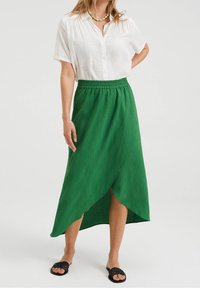 Grüne, strukturierte Wickelröcke mit asymmetrischem Saum, kombiniert mit einem weißen, knöpfbaren Hemd und schwarzen Sandalen.