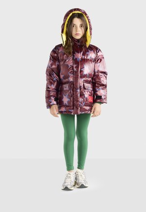 Djevojčica nosi maroon puffer jaknu s uzorkom zvijezda, zelene tajice i bijele tenisice, stoji naslonjena uz jednostavnu svijetlu pozadinu.