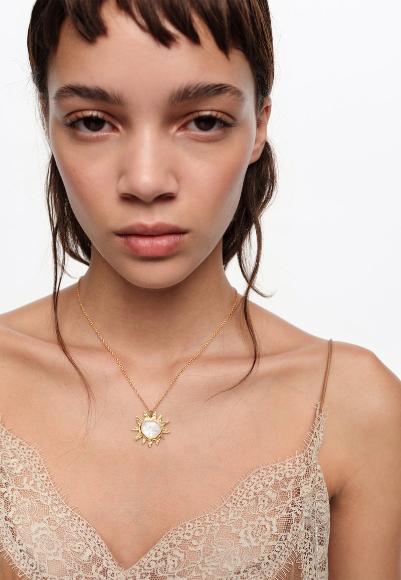 Lühikeste tume juustega naine kannab kuldse päikese kujulist ripatsiga kaelakeed beeži pitsist topi kohal valgel taustal.