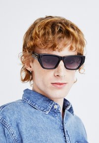 Black sunglasses with a bold geometric design, dark lenses, and gold accents. Worn with a light blue denim shirt. Textured and modern appearance.