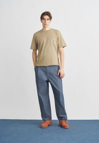 Καφετί βαμβακερό μπλουζάκι με τσέπη και λογότυπο, συνδυασμένο με άνετο μπλε τζιν παντελόνι και πορτοκαλί παπούτσια με κορδόνια και υφασμένη σόλα.