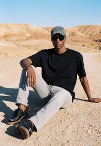 Homme portant une chemise noire, un pantalon gris, une casquette et des lunettes de soleil, assis sur un sol sec et sablonneux avec des collines désertiques en arrière-plan sous un ciel bleu clair.