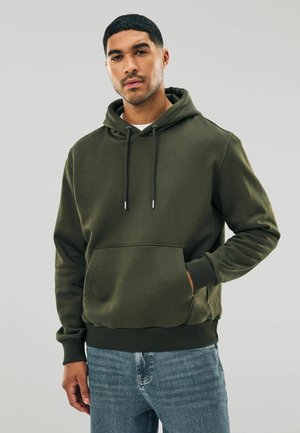 Man som bär en mörkgrön hoodie med framficka och blå jeans, stående mot en ljus, enkel bakgrund.
