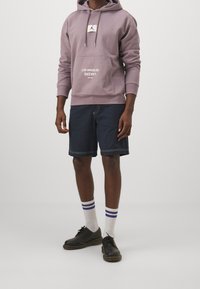 Felpa viola con tasca frontale, logo grafico, abbinata a pantaloni corti di jeans scuri, calze a righe bianche e blu e scarpe nere.