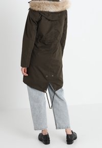 Parka verde oliva con capucha de piel sintética, cintura ajustable y dobladillo curvado. Combinado con jeans de pierna recta azul claro y zapatos negros.