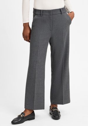 Person iført grå vidde ben bukser og sorte loafers med gyldne spænder, stående med den ene hånd i lommen mod hvid baggrund.