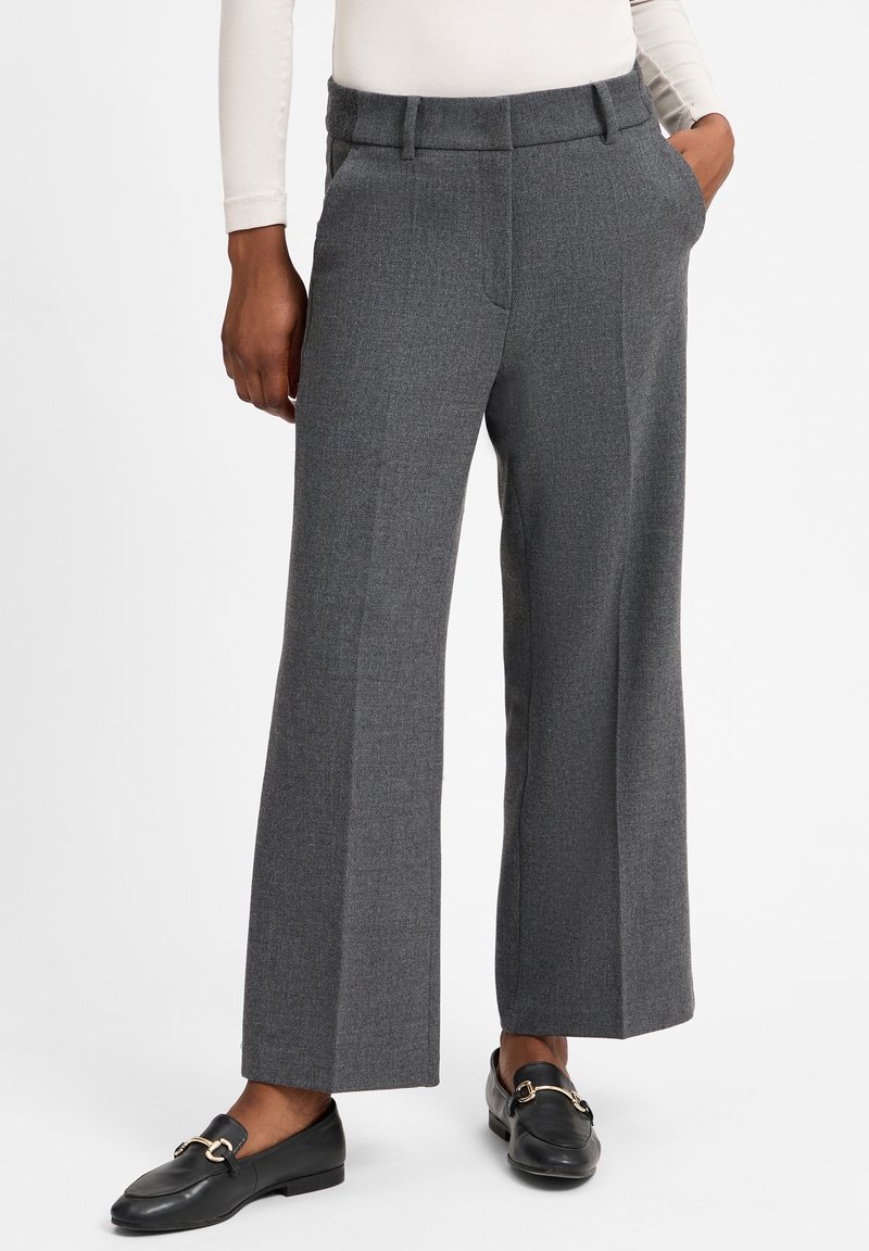 Personne portant un pantalon gris à jambes larges et des mocassins noirs avec des boucles dorées, debout avec une main dans la poche sur fond blanc.