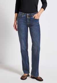Jeans en denim bleu à coupe droite, dotés d'une braguette à boutons, d'un design à cinq poches et de détails de couture visibles. Portés avec des chaussures en mesh noires.