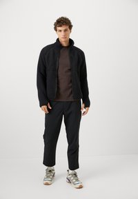 Giovane in piedi che indossa una giacca in pile nera, una camicia marrone scuro, pantaloni neri, calzini bianchi e scarpe da trekking beige con accenti blu.