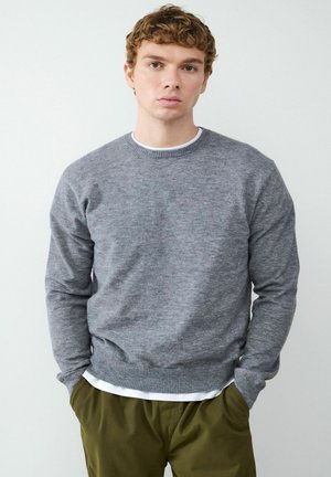 Grau geschnittener Pullover aus weichem Strickmaterial. Verfügt über gerippte Bündchen und Saum, wird über einem weißen T-Shirt getragen und mit olivgrünen Hosen kombiniert.