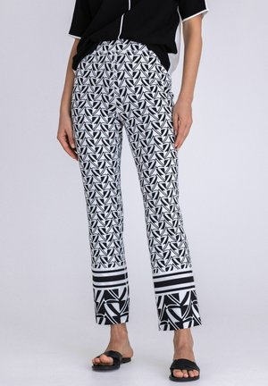 Woman wearing black and white geometric patterned pants with striped cuffs, black top, and black flat sandals, standing against plain background.