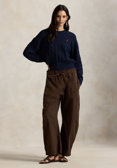 Μπλε πουλόβερ με πλέξη καλώδιο και φαρδιά μανίκια, συνδυασμένο με χαλαρά καφέ παντελόνια και καφέ σανδάλια. Διαθέτει λογότυπο στο πουλόβερ.