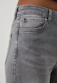 Gros plan d'une personne portant un jean en denim gris délavé montrant les détails de la poche avant et de la ceinture sur un fond neutre.