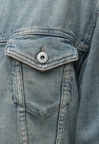 Ανοιχτό μπλε τζιν μπουφάν με τσέπη στο στήθος, που διαθέτει ασημί κουμπί με χαραγμένο το "HUGO JEANS", ορατές ραφές και υφή ύφανσης.