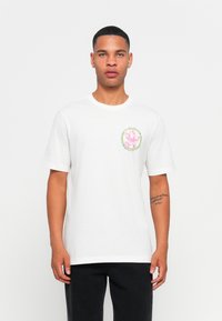 Jovem com cabelo curto, vestindo uma T-shirt branca com uma pequena gráfica colorida no lado esquerdo do peito e calças pretas, em pé contra um fundo simples.