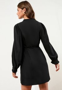 Schwarzes Kleid mit figurbetontem Schnitt, langen Puffärmeln und Rüschenmanschetten. Glatter Stoff mit hochgeschlossener Halslinie und Taillendetail.