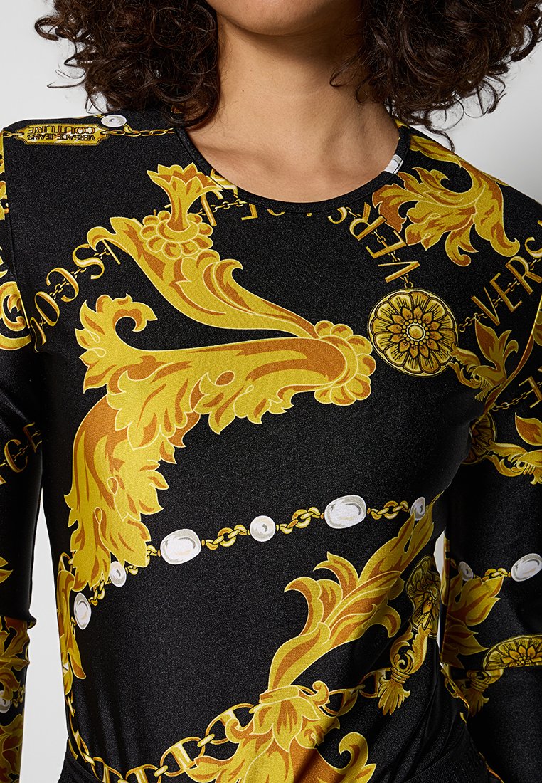 Haut à manches longues noir présentant des motifs floraux dorés intricats et des accents de perles blanches, fabriqué dans un tissu lisse et légèrement brillant.