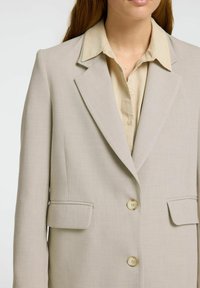 Blazer ajustado de color beige claro con solapas de muesca, dos bolsillos frontales y dos botones. Lleva puesto una camisa de color claro con cuello.