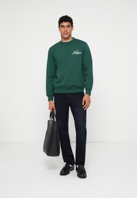 Grønn genser med rund hals og hvit logo, kombinert med marineblå bukser og svarte sko. Bærer en svart totebag.