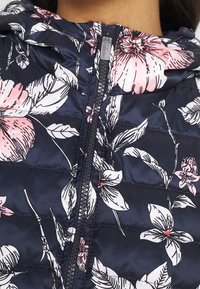 Laivastonsininen kukkakuosisella toppatakkilla, jossa on vaaleanpunaisia ja valkoisia kukkakuvioita, tikattu muotoilu, etuvetoketju ja säädettävä huppu. Sileä, kiiltävä kangas.