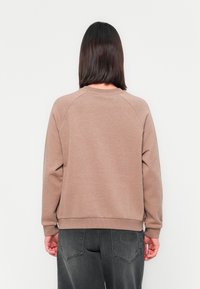 Personne aux cheveux foncés mi-longs de dos, portant un sweat-shirt marron clair et un jean gris foncé avec poches arrière visibles.