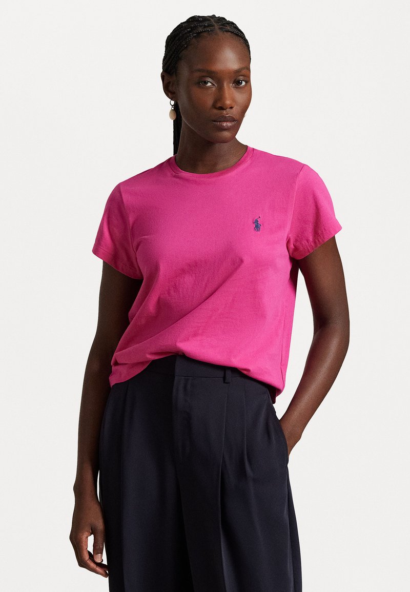 Rosa Baumwoll-T-Shirt mit kurzen Ärmeln, rundem Halsausschnitt und einem kleinen blauen Logo auf der Brust, kombiniert mit dunklen Hosen.