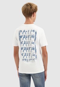 Jongeman met kort, lichtbruin haar, gekleed in een wit T-shirt met een herhaald blauw "BALLIN"-ontwerp op de achterkant en een blauwe spijkerbroek.