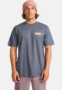 Camiseta de mangas cortas azul grisácea con cuello redondo y pequeño parche con logo en el pecho. Combinada con pantalones de pana en rosa claro.