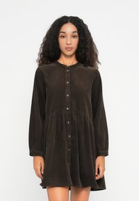 Donna con capelli ricci che indossa un vestito di velluto a coste marrone scuro con maniche lunghe e bottoni frontali, in piedi di fronte a uno sfondo chiaro e uniforme.