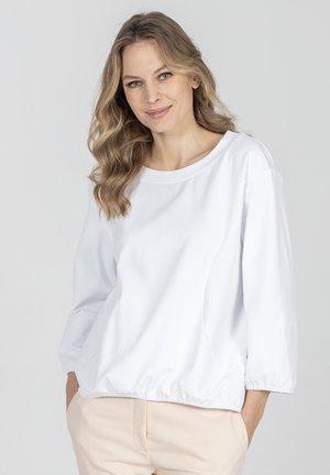 Kobieta z falującymi blond włosami, ubrana w luźną białą koszulę z długim rękawem i jasnobeżowe spodnie, uśmiechająca się z rękami w kieszeniach, na jednolitym szarym tle.