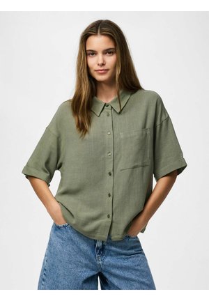 Kobieta z długimi włosami, ubrana w luźną oliwkowozieloną koszulę na guziki z podwiniętymi rękawami i niebieskie jeansy, ręce w kieszeniach, na białym tle.
