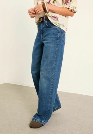 Kvinde iført blå vidde jeans, brun bælte, blomstrede bluse og brune sko, stående på beige gulv mod en hvid væg.