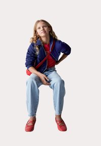 Chaqueta bomber marina, bolso cruzado rojo, camisa roja, vaqueros holgados de un azul claro y bailarinas rojas con tiras entrelazadas, sentada contra un fondo neutro.