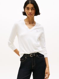 Hvit V-hals genser laget av mykt materiale, tettsittende design, brukt med mørkeblå jeans og et svart belte med en gullspenne som akcent.