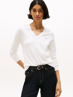 Hvid V-hals trøje lavet af blødt materiale, tætsiddende design, båret med mørkeblå jeans og et sort bælte med en guldspændeaccent.