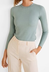 Maglietta a maniche lunghe a costine di un verde tenue, con scollatura a girocollo. Abbinata a pantaloni beige chiaro su misura, creando una silhouette slanciata.