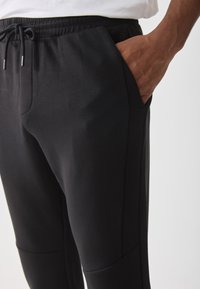 Svart joggebukse laget av mykt stoff med elastisk linning, sidelommer og en smalere ben design. Jevn tekstur med minimale sømmer.