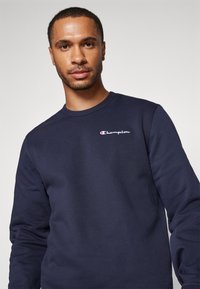 Sweat-shirt col rond bleu marine en tissu doux, avec un petit logo Champion brodé sur la poitrine. Manches longues avec une coupe décontractée.