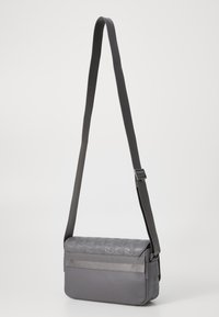 Šedá crossbody taška s texturovaným vrchem a hladkým spodkem, vybavená širokým nastavitelným popruhem a nenápadným kovovým akcentem.