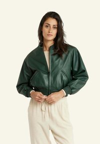 Veste bombardier en cuir vert avec des poignets et un ourlet côtelés, dotée de poches latérales et d'une fermeture éclair. Portée sur un haut et un pantalon beige neutres.