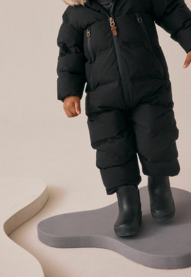 Tout-petit portant une combinaison d'hiver noire matelassée et des bottes noires, debout sur une plateforme grise irrégulière sur fond beige.