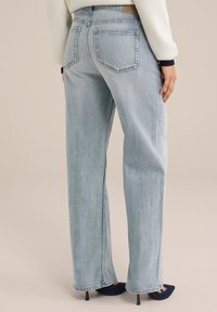 Jeans de cintura alta en azul claro, con pierna ancha, que cuentan con dos bolsillos traseros y una textura desgastada. Combinados con tacones azul marino.