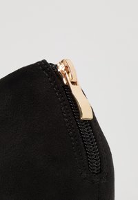 Černé semišové kotníkové boty se zlatým zipem. Zip se táhne po boku a doplňuje hladkou strukturu materiálu.