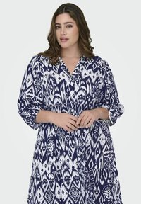 Mornarska in bela obleka s vzorcem z V-izrezom, zavihanimi rokavi in večplastnim dizajnom. Izdelana iz gladkega blaga z drzno ikatno potiskom.