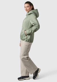 Haut de survêtement léger de couleur vert clair, avec une fermeture éclair à l'avant, des poches latérales et une capuche ajustable. Porté avec un pantalon beige et des chaussures de sport noires.