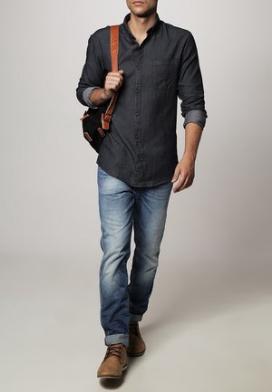 Camicia button-up grigia scura da uomo con taschino, abbinata a jeans sbiaditi blu chiaro e scarpe marroni con lacci, mentre porta una borsa nera.