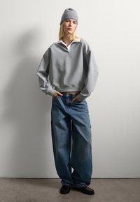 Grijze cropped sweatshirt met een witte kraag, gecombineerd met loszittende blauwe jeans en zwarte schoenen. Model staat tegen een effen achtergrond.